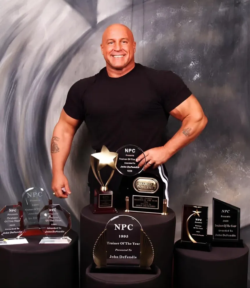 Smiling man in black shirt stands behind award plaques and trophies on round tables, with a gray backdrop.
