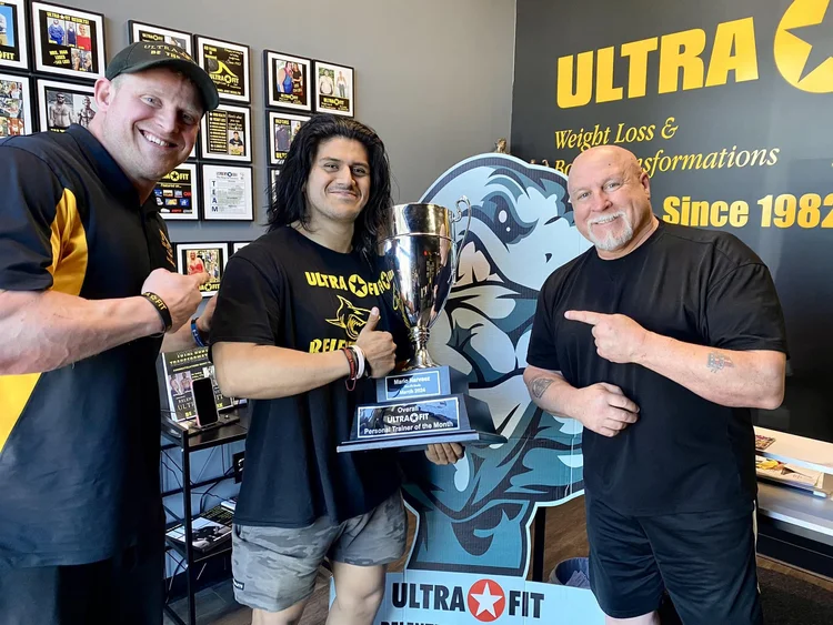 Three men smiling indoors, with one holding a trophy; gym awards and logos visible in the background.