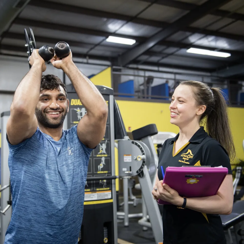 Man lifting dumbbells overhead while a woman with a clipboard smiles and observes in a gym setting.