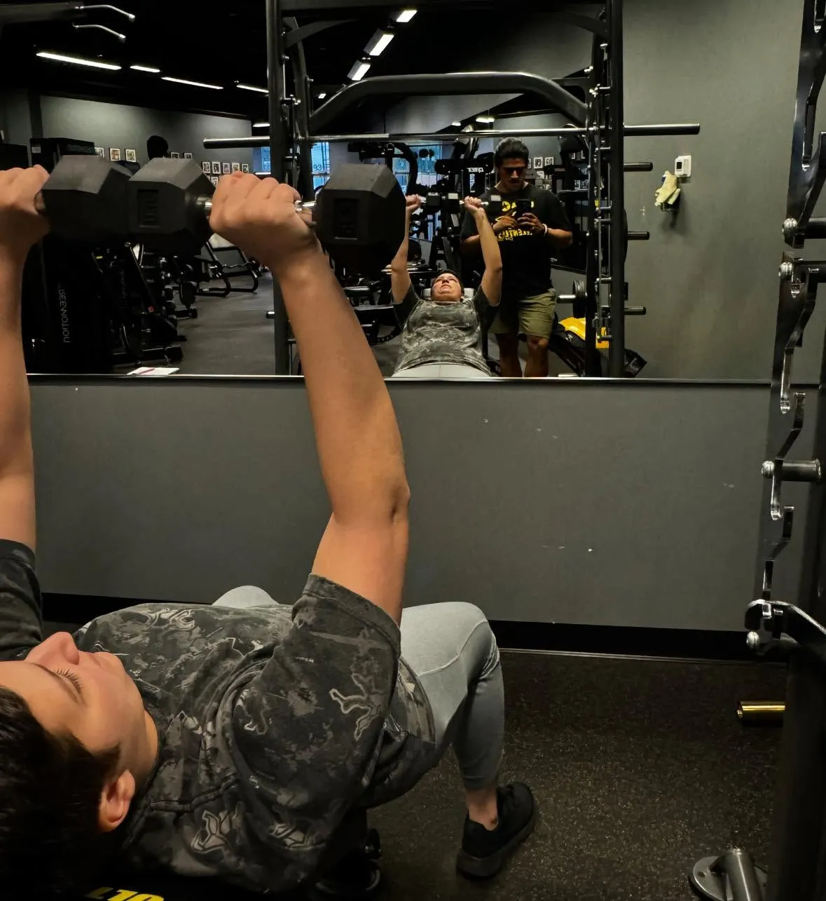 Person doing dumbbell bench press on a gym bench, reflected in a mirror with another person taking a photo.