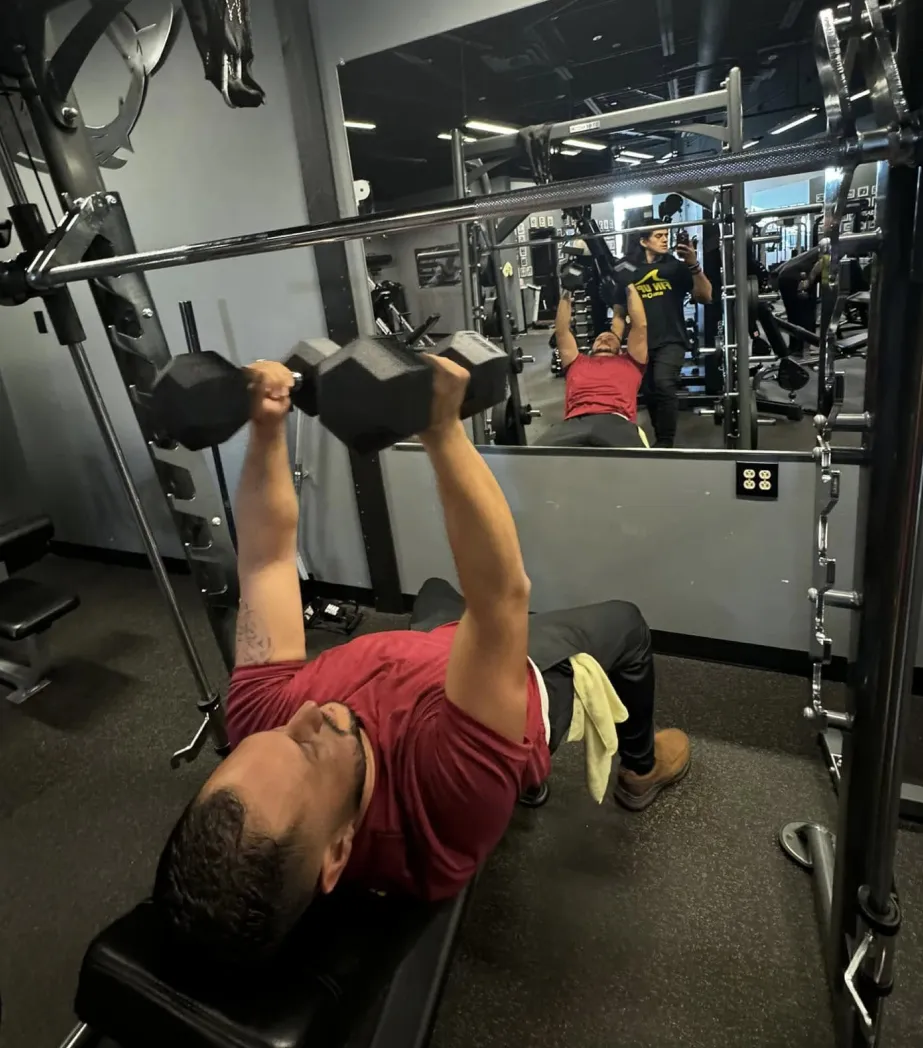 Man in red shirt doing dumbbell bench press on a bench at the gym, mirror reflecting his workout.