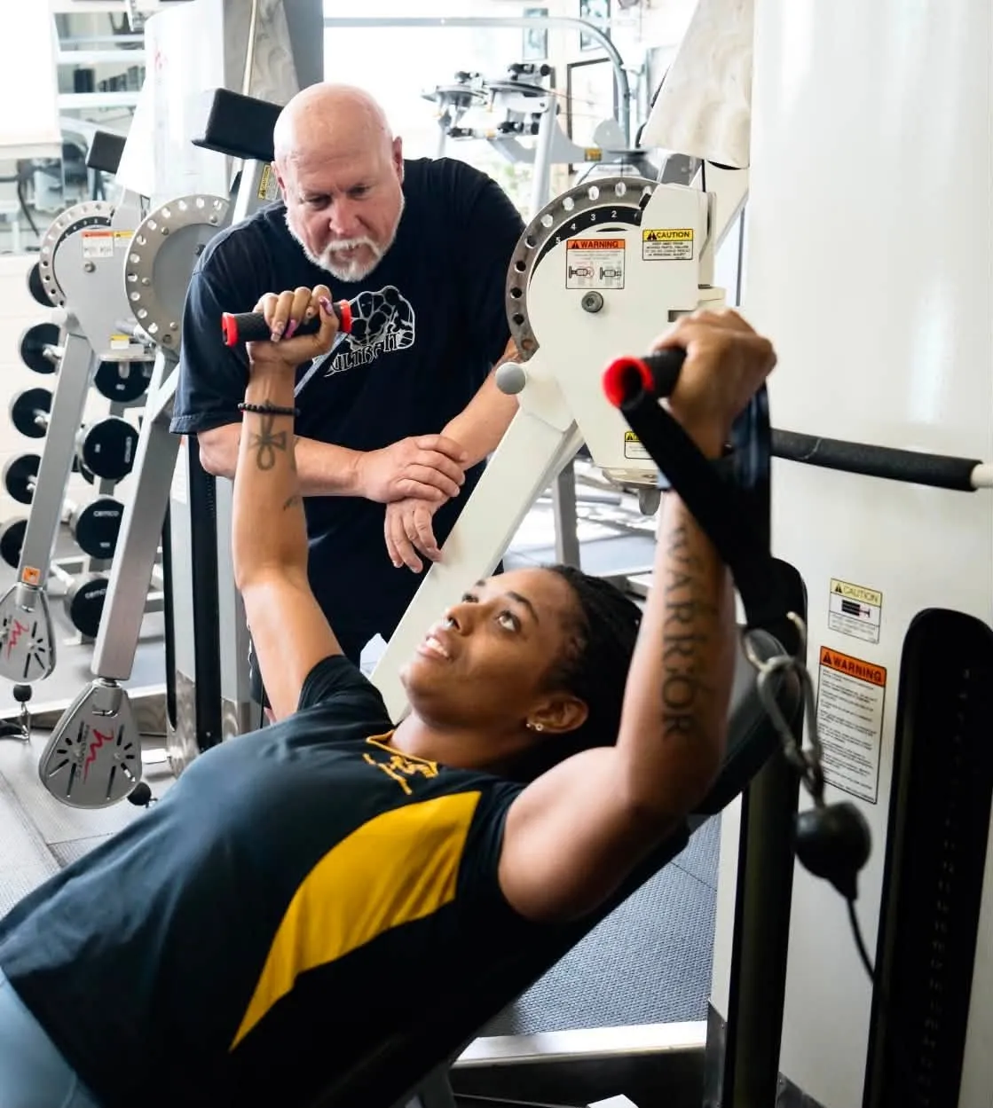 A person lifts weights on a machine while a trainer observes and coaches them in a gym setting.
