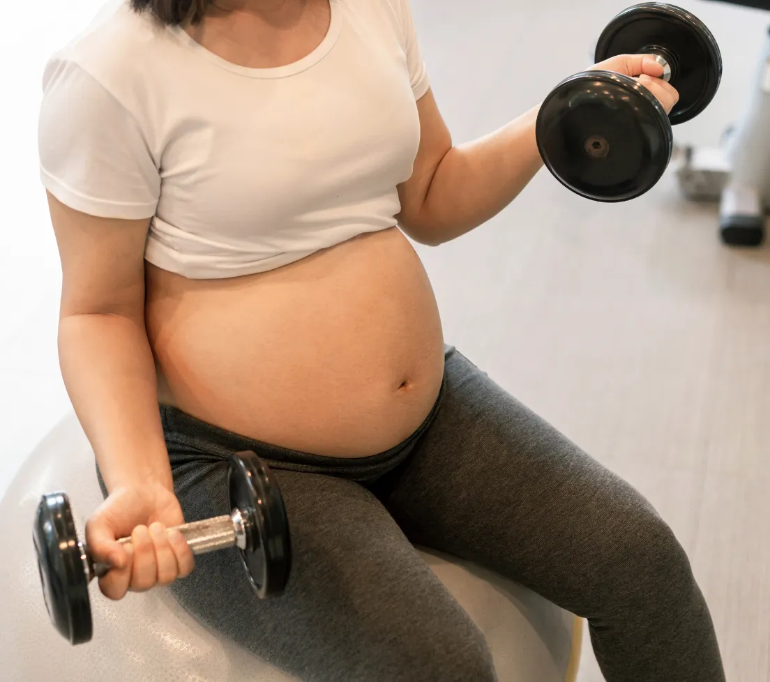 Pregnant woman in workout clothes sitting on a ball and lifting dumbbells in both hands.