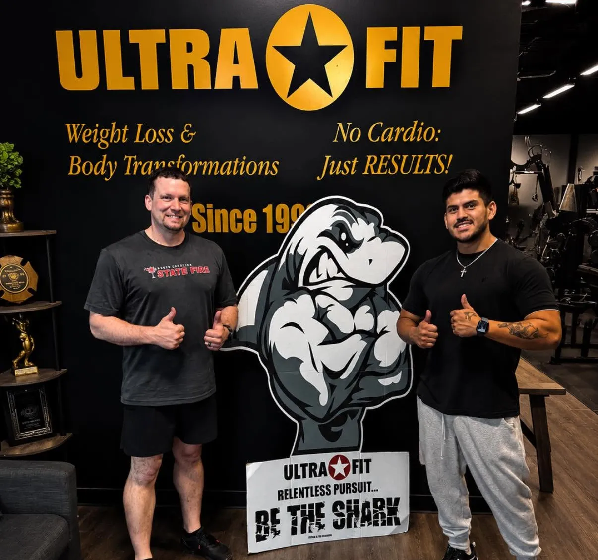 Two men stand in a gym by a shark logo, smiling and giving thumbs up in front of an Ultra Fit sign.