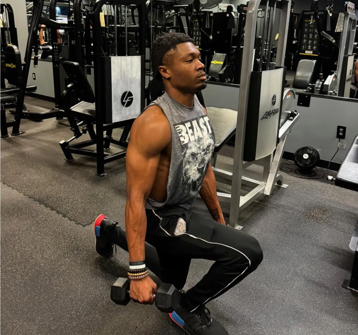 A man performs a lunge with dumbbells in a gym, wearing a sleeveless shirt and black workout pants.