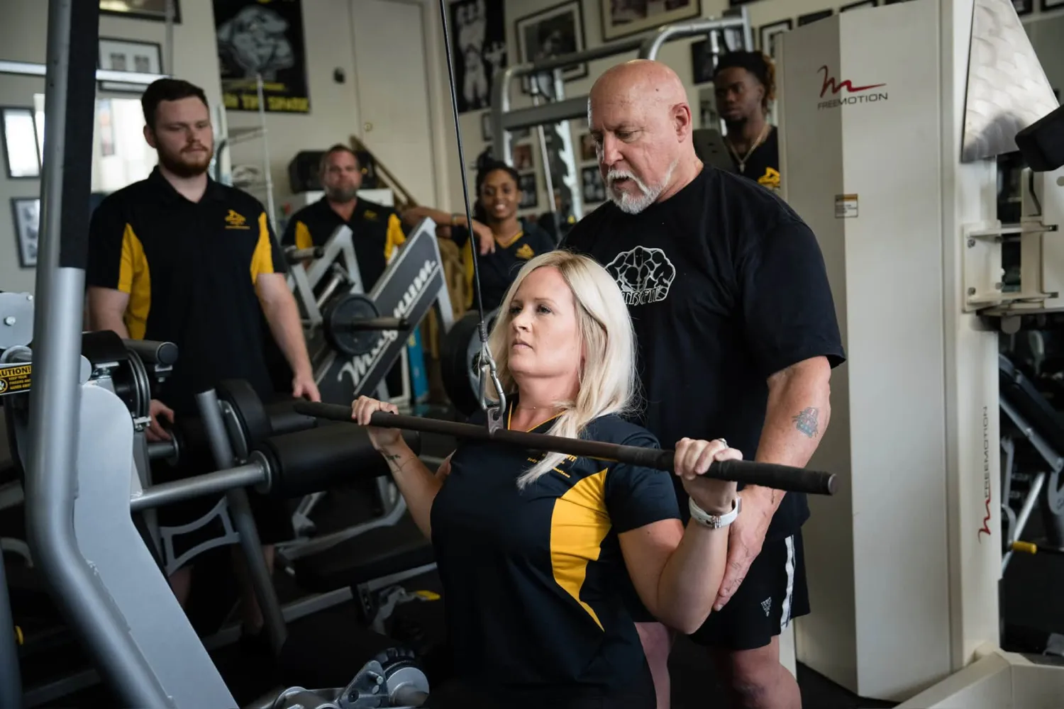 A woman lifts weights at the gym with a trainer spotting her; several others watch in the background.