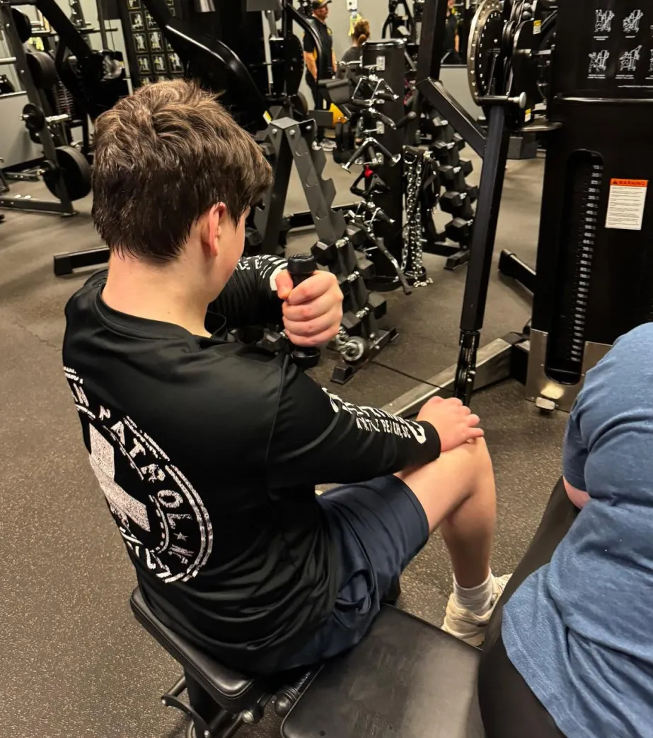 A person in a black shirt sits on a bench in a gym, resting their elbow on their knee near weight machines.