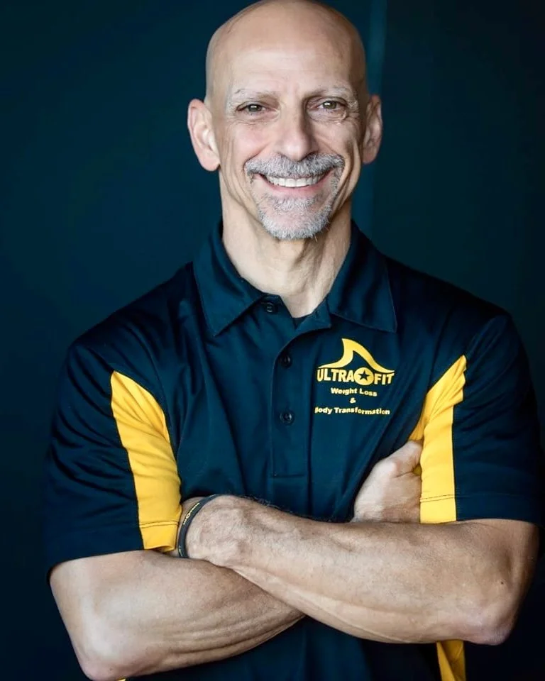 Smiling bald man in a black and yellow “Ultra Fit” shirt stands with arms crossed against a dark background.