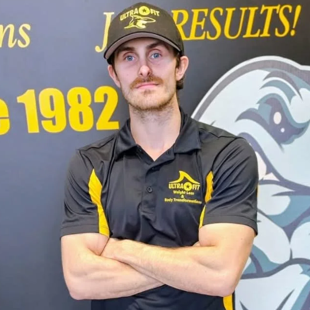 Man in a black and yellow “Ultra Fit” uniform stands with arms crossed in front of a gym-themed backdrop.