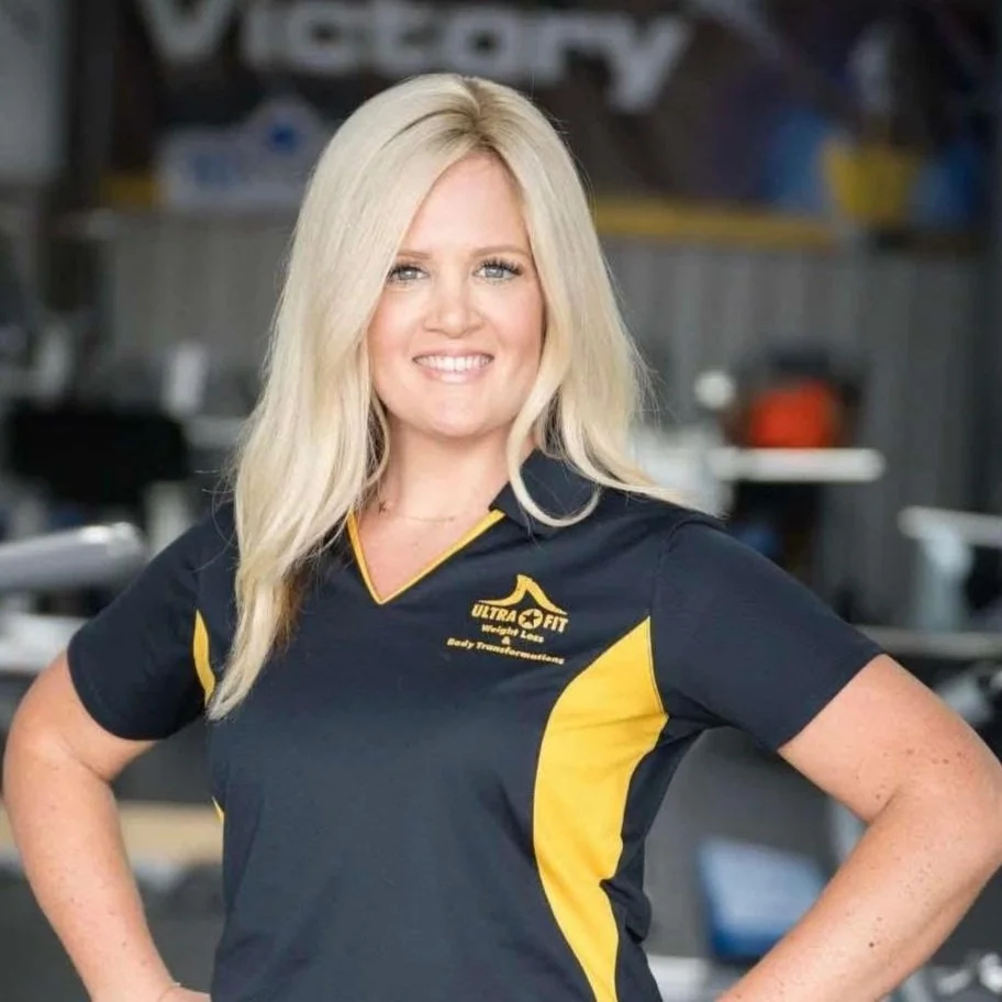 Smiling woman with long blonde hair in a navy and yellow polo shirt stands with hands on hips indoors.