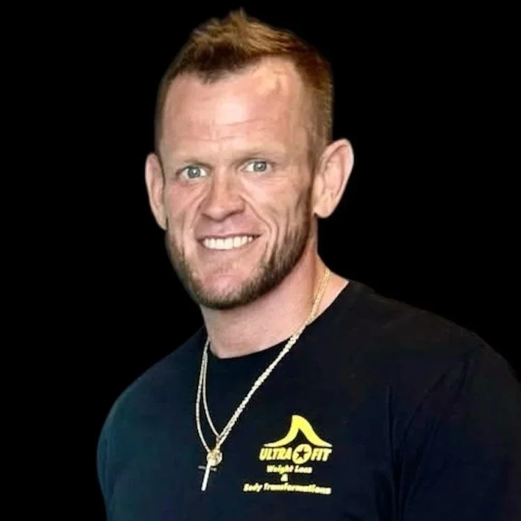 Smiling man with short hair in a black t-shirt and gold necklace on a plain black background.
