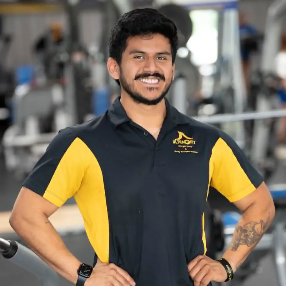 Man in a black and yellow polo shirt smiling in a gym, standing with hands on hips.