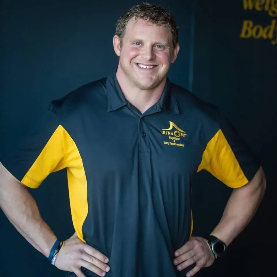 Man in a black and yellow polo shirt smiles with hands on hips, standing against a dark background.